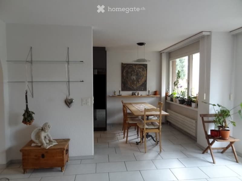 Bright, minimalist dining area with a wooden dining table and chairs, a small wooden stool with a decorative sculpture, potted plants on a side table, and a wall-mounted art piece.