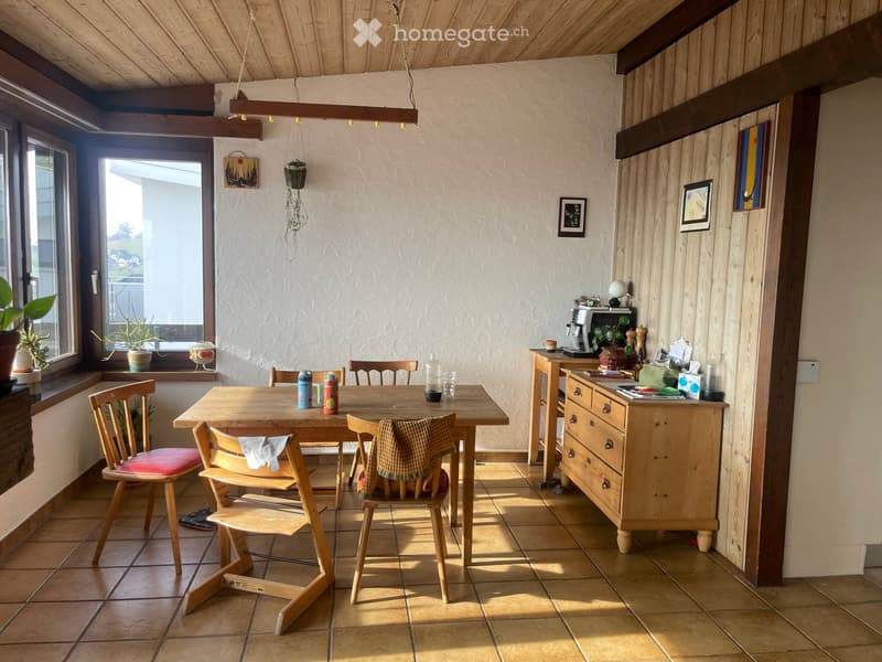 Bright kitchen with wooden table and chairs, tiled floor, and wooden cabinetry. Features a coffee maker, fridge, and various kitchen items on the counter.