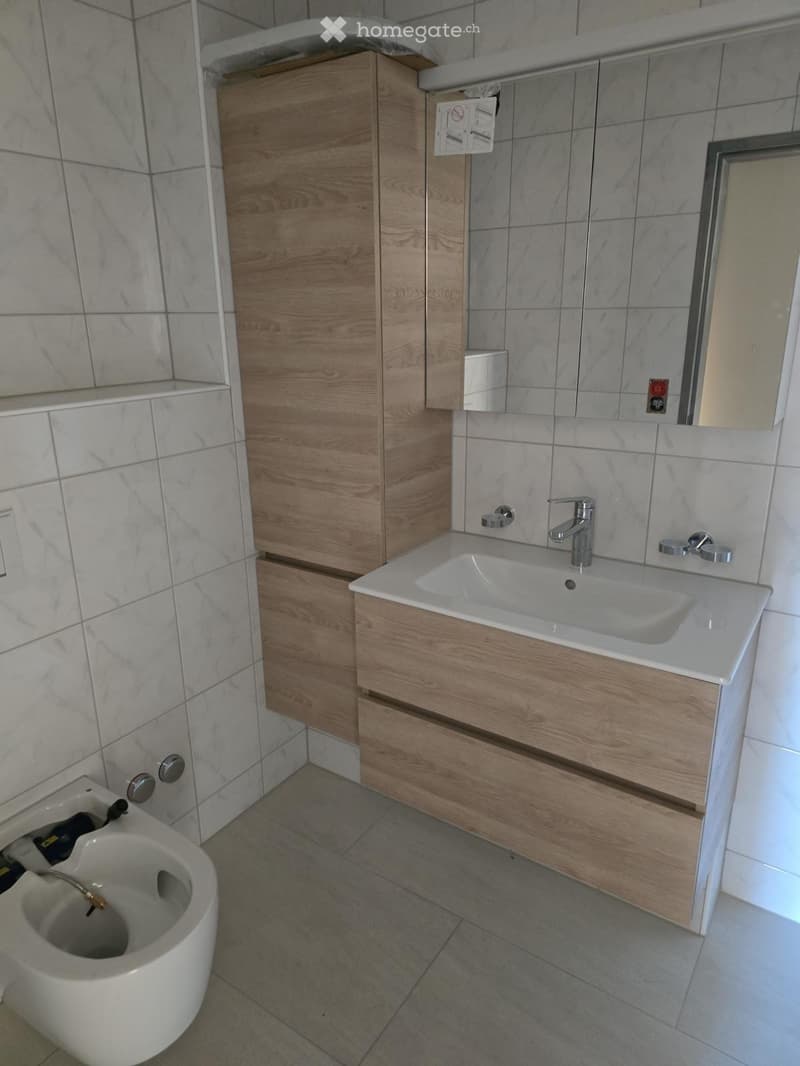 Modern bathroom with a white sink, wooden cabinet, and a toilet. The walls are tiled with marble-like patterns, and the floor is tiled with gray tiles.