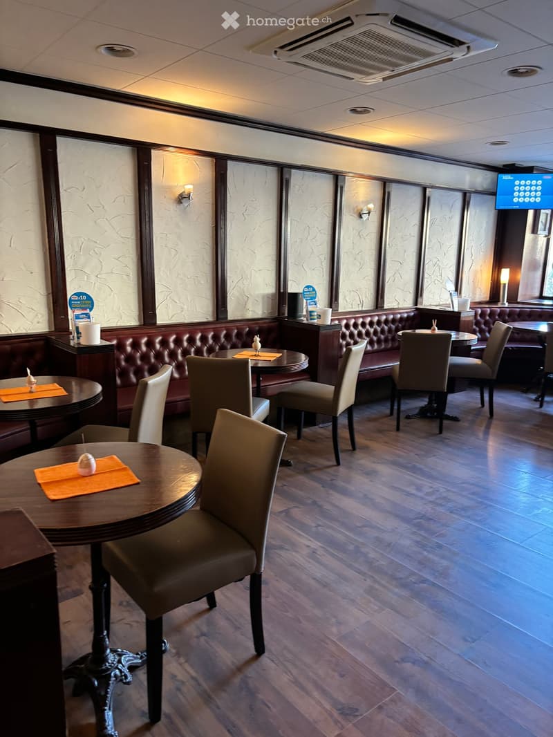 Elegant restaurant interior with wooden floors, round tables, upholstered chairs, and a long leather sofa along the wall.