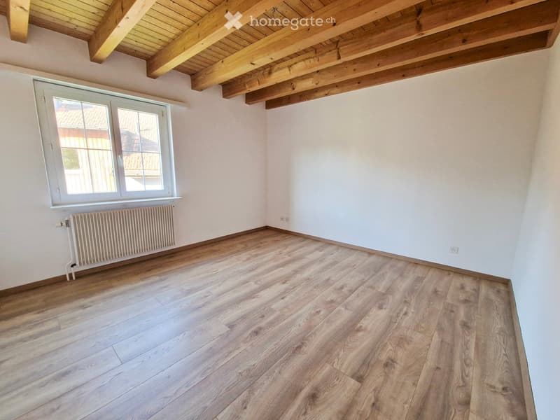Empty room with wooden flooring, white walls, exposed wooden ceiling beams, and a single window with white frame. Radiator is visible under the window.