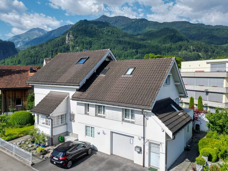 A two-story white house with a brown tiled roof, multiple windows, and a garage. A black car is parked in front of the garage. The house is surrounded by greenery and is situated in a scenic area with mountains in the background.