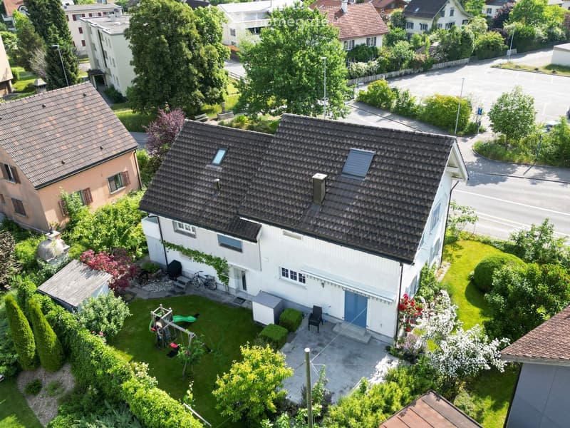 Aerial view of a two-story house with a well-maintained garden, featuring a patio, swing set, and surrounding greenery.