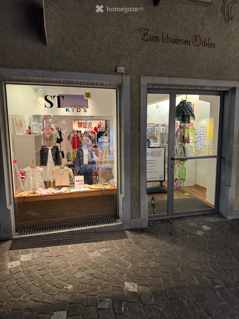 Storefront with glass doors, display of children's clothing, sign indicating sale, and a lamp outside