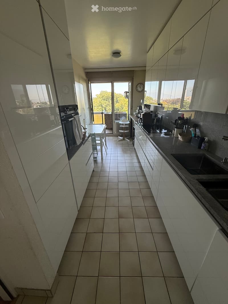 Modern kitchen with white tiled floor, light-colored cabinets, and a black countertop. Features include a sink, oven, small table with chairs, and a clock on the wall. Large windows and a sliding glass door lead to a balcony.