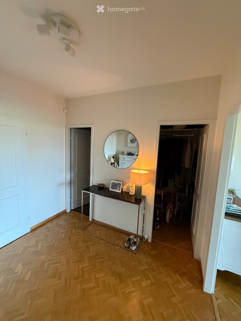 Hallway with herringbone wooden flooring, a small black table with a lamp, a mirror, and a picture frame. A closet with clothes and bicycles is visible in the background.