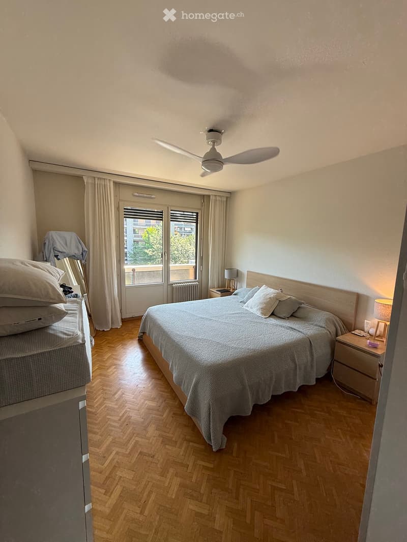 Cozy bedroom with wooden herringbone flooring, large double bed with pillows and blankets, nightstand with lamp, ceiling fan, and large window with curtains leading to a balcony.