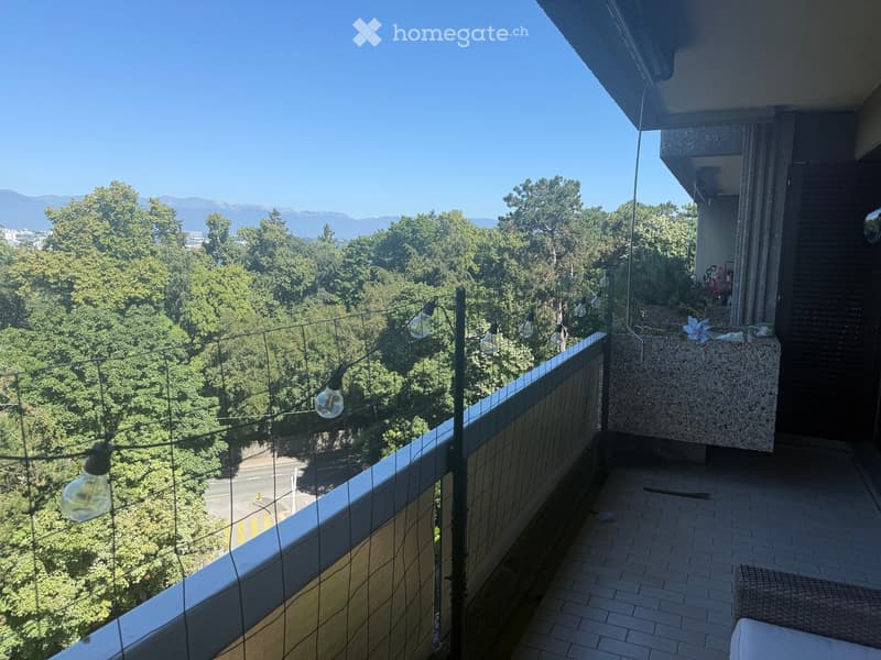 Balcony with a view of trees and mountains, featuring a glass railing, string lights, and a small seating area with a wicker chair and table.