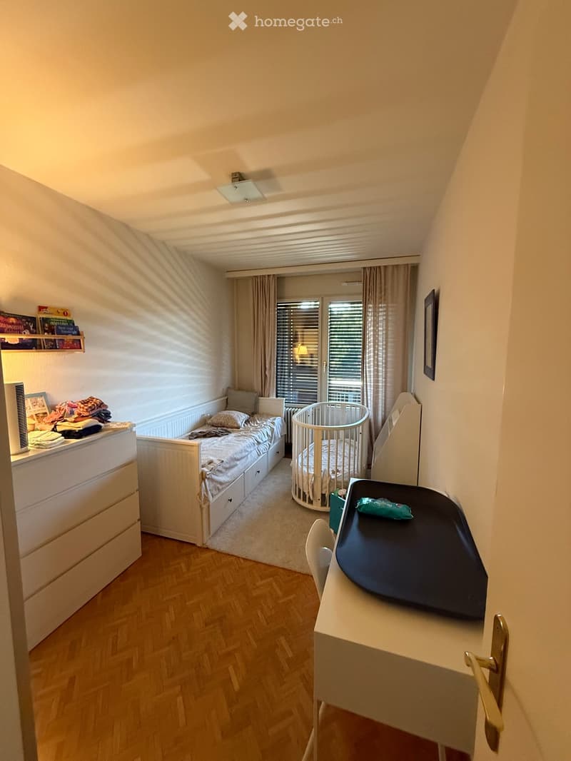 Cozy bedroom with a single bed, baby crib, changing table, and a shelf with books and folded clothes. The room features wooden flooring and large windows with curtains.