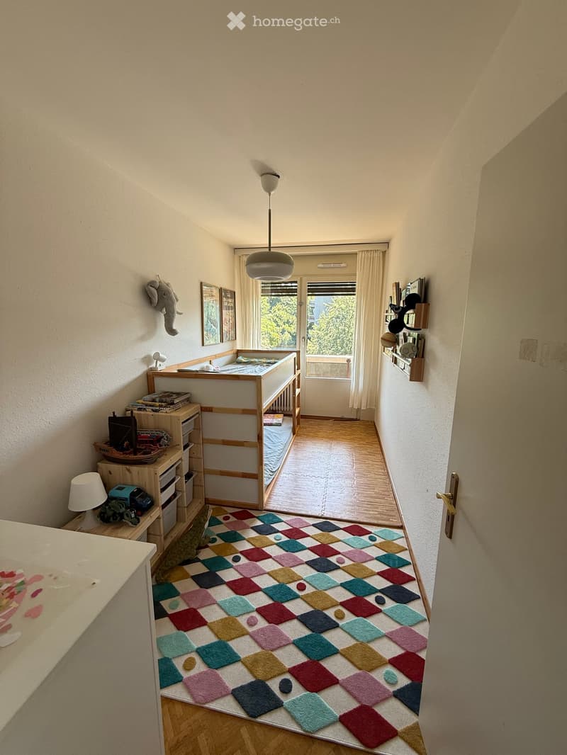 A cozy children's bedroom with a colorful rug, a bunk bed, a small table, and a storage unit. The room features a large window with curtains and a view of greenery outside.