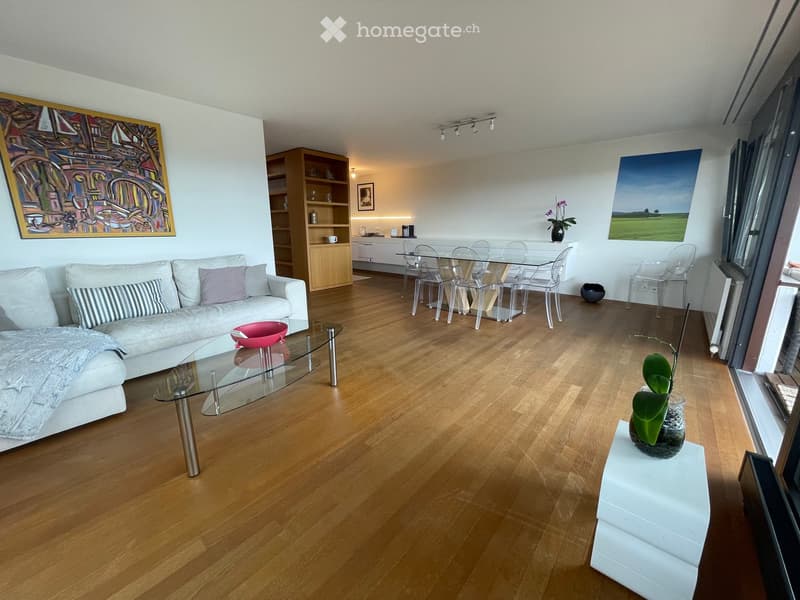 Modern living room with light wooden flooring, white sofa, glass coffee table, dining area with glass table and chairs, and decorative artwork on the walls.