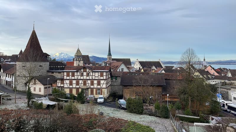 A picturesque town with traditional houses, a church tower, and snow-capped mountains in the background.