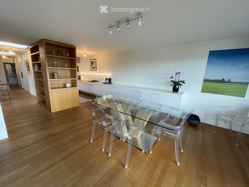 Modern dining area with a glass table and transparent chairs, a kitchen counter, and a wooden cupboard with shelves.