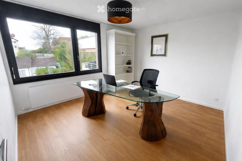 Modern office space with a glass desk, office chair, laptop, bookshelf, and a large window offering a view of the outdoors.