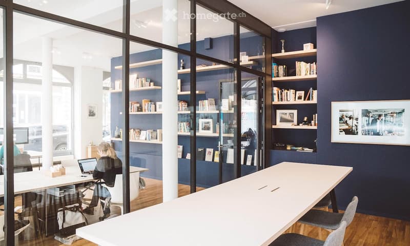 Modern office space with dark blue walls, white tables, chairs, shelves filled with books and decorative items, and a laptop on a desk.