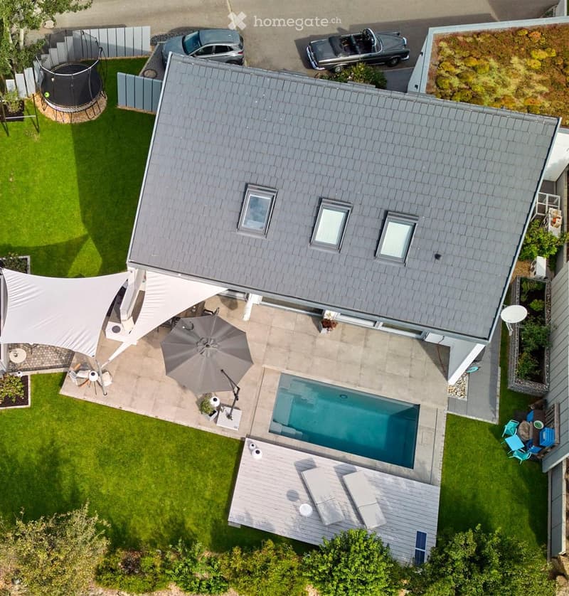 Aerial view of a modern house with a swimming pool, patio, outdoor furniture, and a trampoline in the yard.