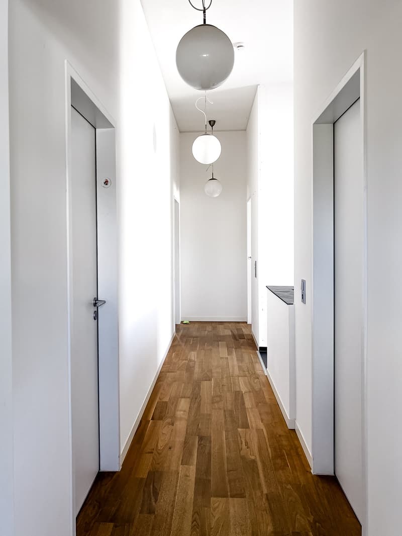 A narrow hallway with white walls, wooden flooring, and modern pendant lights hanging from the ceiling. The hallway features several doors and a small built-in shelf on the right side.