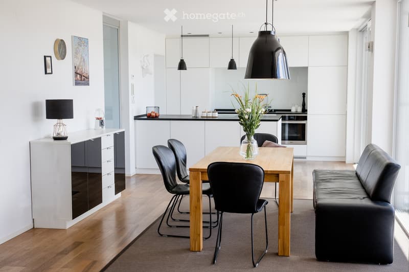 Modern open-plan living and dining area with a wooden dining table, black leather chairs, a black leather sofa, pendant lights, and a kitchen area with white cabinets and a black countertop.