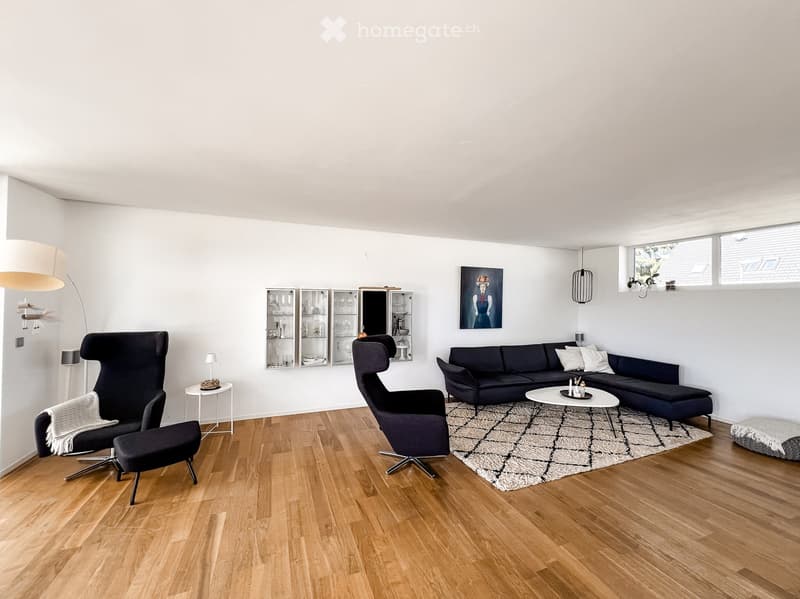 Modern living room with light wooden flooring, white walls, and large windows. Features a dark blue sectional sofa, coffee table, armchairs, and a patterned rug.