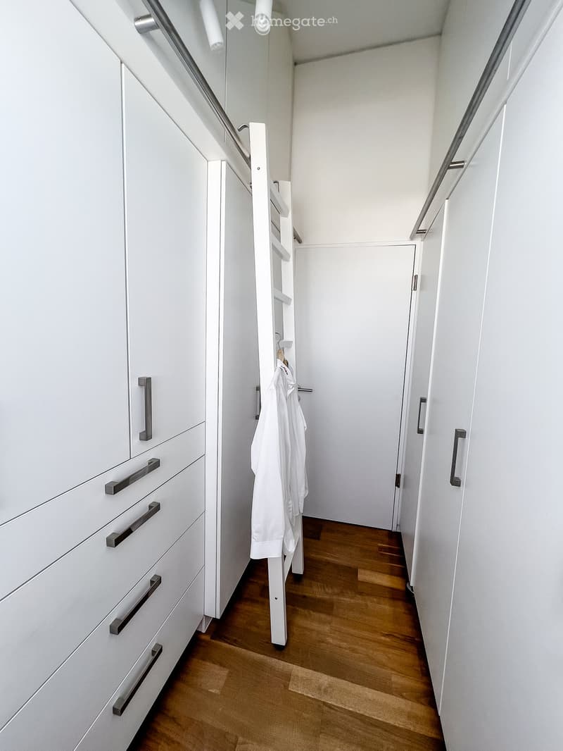 Modern hallway with white walls, wooden flooring, built-in white cabinets with black handles, and a small coat rack holding white garments.