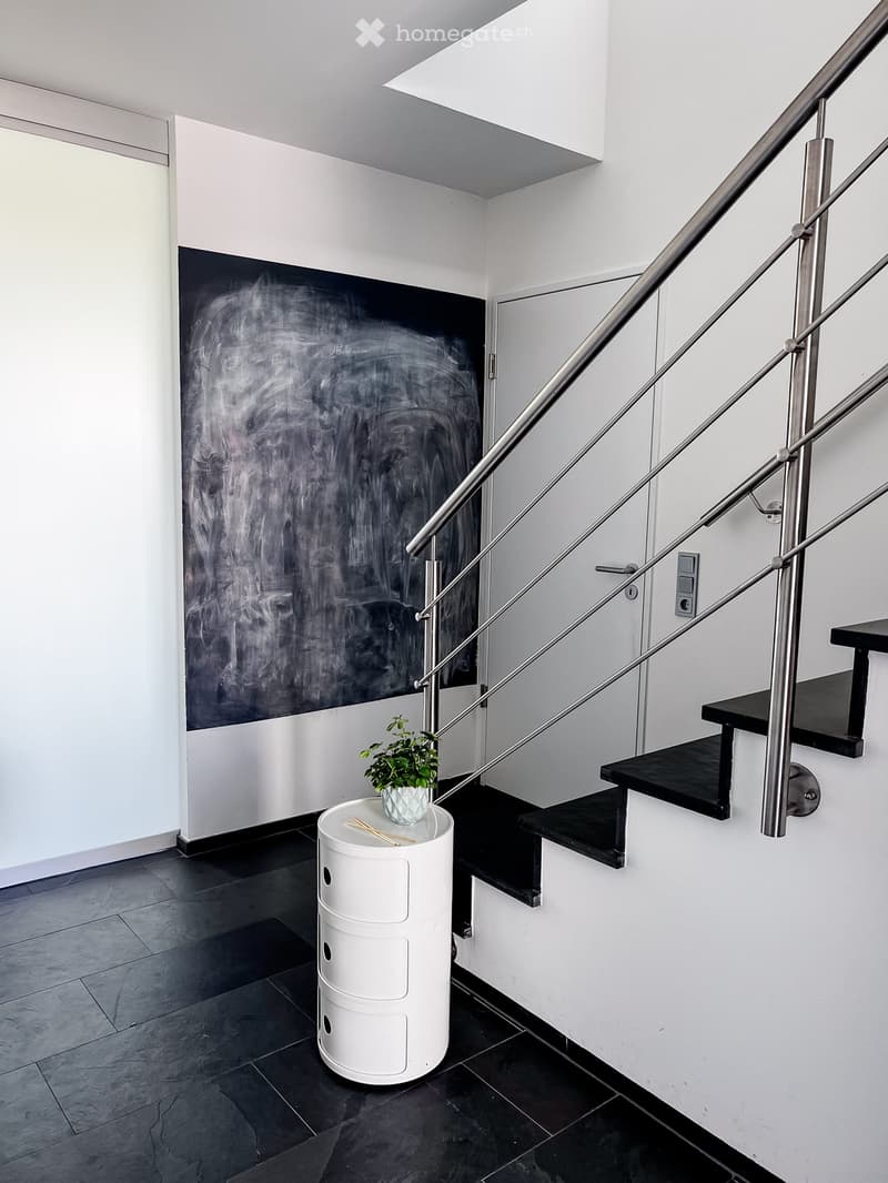 Modern hallway with a sleek staircase featuring a stainless steel railing, black steps, and a white cylindrical storage unit with a small potted plant on top.