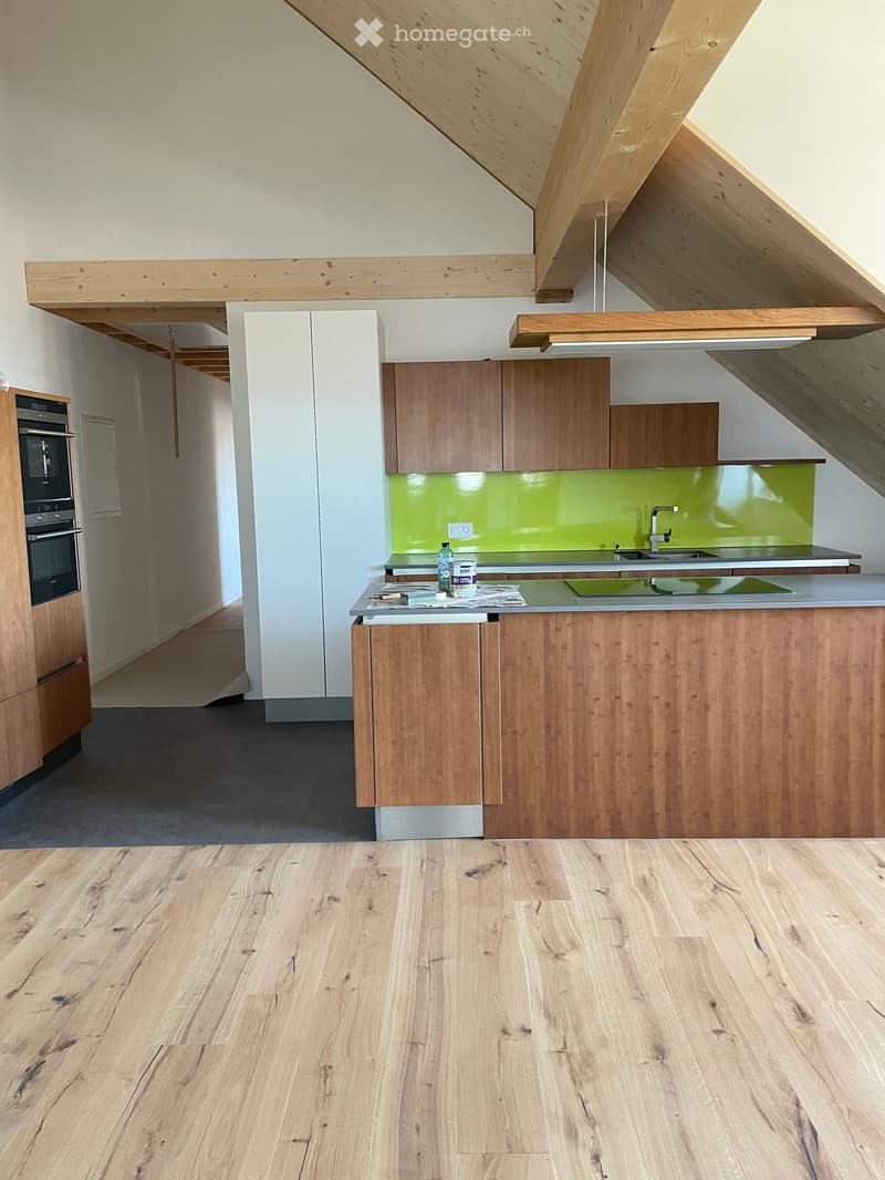 Modern kitchen with wooden cabinets, a central island, and a green backsplash. The floor is light wood, and the ceiling features exposed wooden beams.