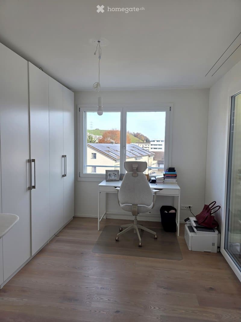 A modern, minimalist office space with a wooden floor, white walls, large windows, a grey office chair, a white desk, and a few decorative items.