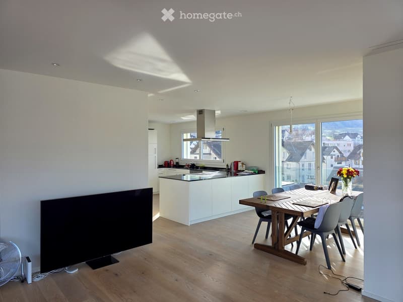 Open-plan living and dining area with a large wooden dining table, modern chairs, a kitchen area with white cabinets, and a large TV screen on the wall.