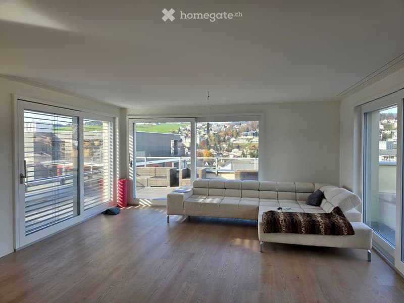Modern living room with light-colored sectional sofa, coffee table, ottoman, and large windows offering a scenic view. The room features wooden flooring and a balcony accessible through large glass doors.