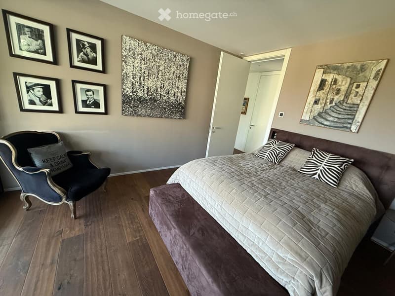 Large bed with grey bedding and pillows, grey upholstered headboard, chair with pillow, wooden flooring, paintings on the wall.