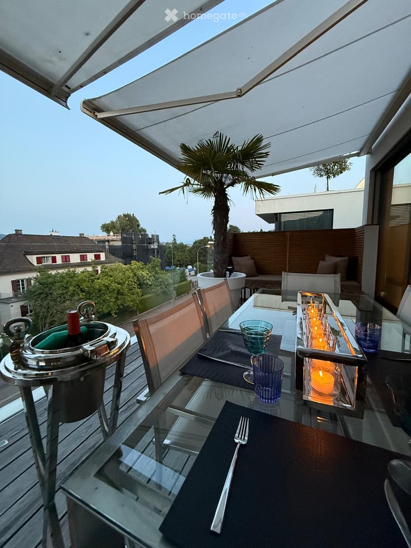 A covered outdoor dining area with a table and chairs, overlooking a cityscape with buildings and trees. A bottle of wine is chilling in a bucket. There is a palm tree in the center. It's a serene and beautiful place for enjoying a meal or drinks in the o