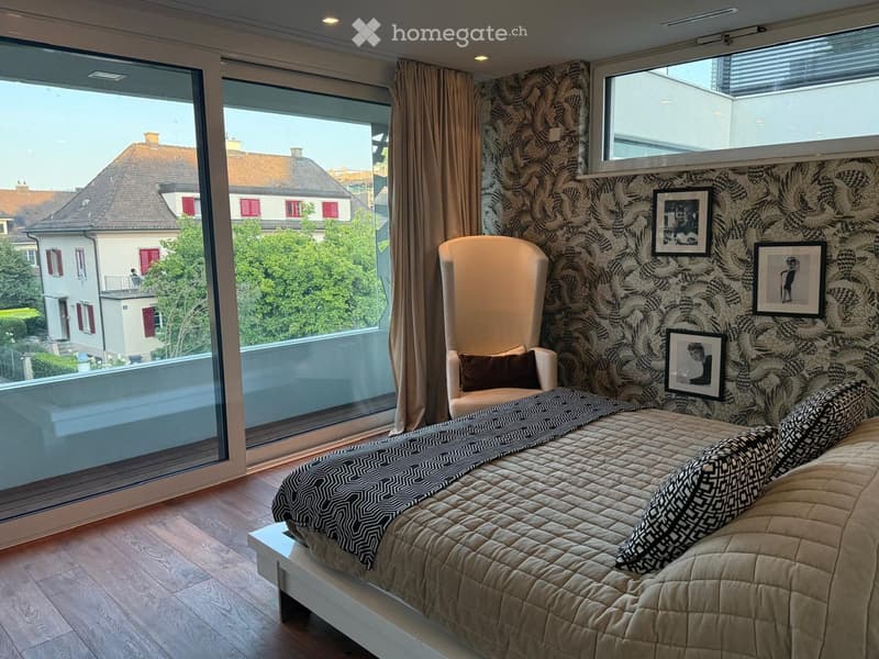Bedroom with double bed, white bed frame, light wood floor, beige quilt, patterned pillows, a white chair with pillow on the side, large glass door to balcony, panoramic window