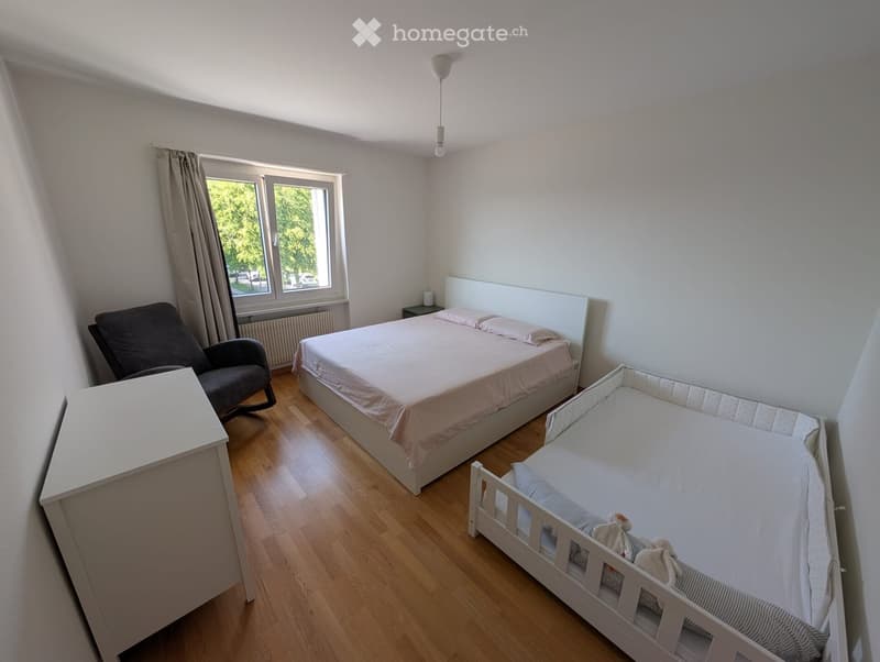 Bright bedroom with wooden flooring, a double bed with pink bedding, a baby crib, a dark armchair, and a white nightstand.