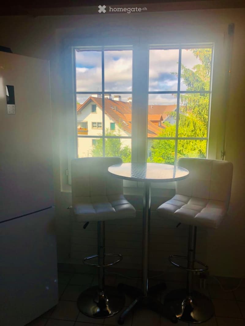Bright room with two white stool chairs and a round table, large windows overlooking a residential area.