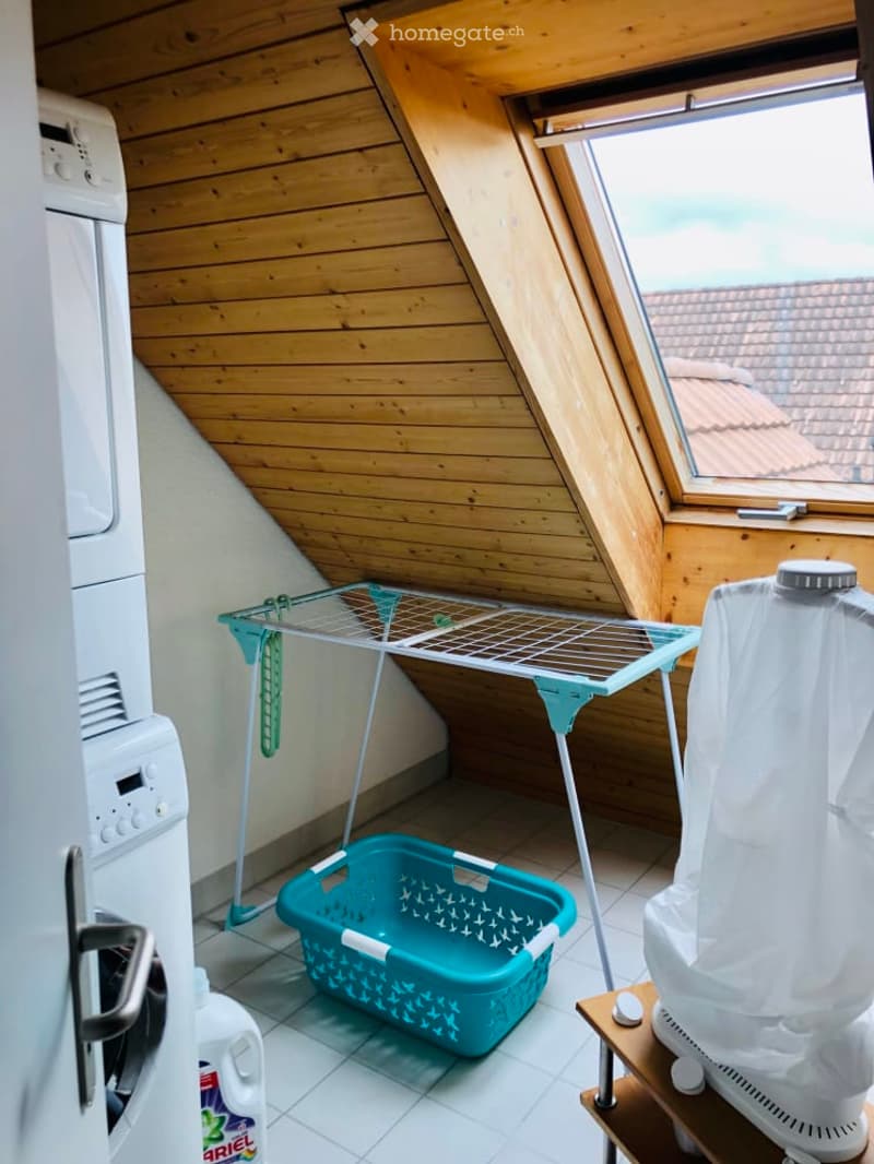 Attic laundry room with wooden slanted ceilings, a washing machine, a drying rack, a laundry basket, and a small stool.