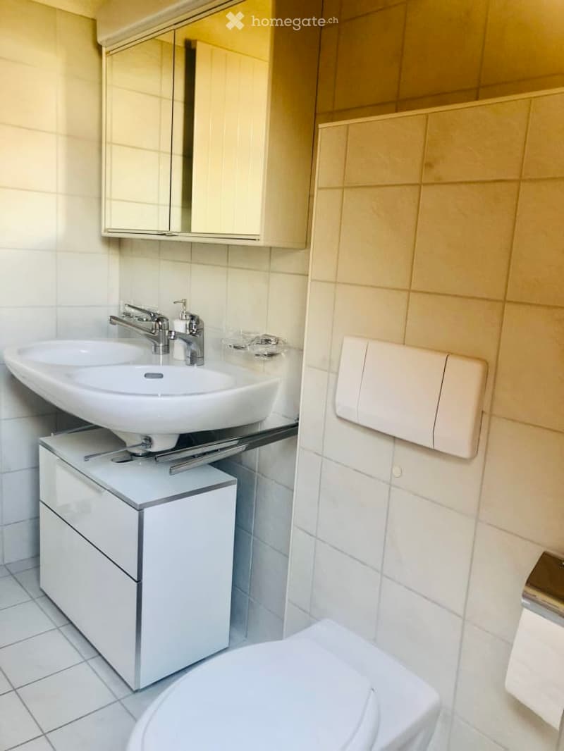 Modern bathroom with a white sink, chrome faucet, mirrored cabinet, and a toilet. The walls are tiled in light beige, and the floor is white tiled.