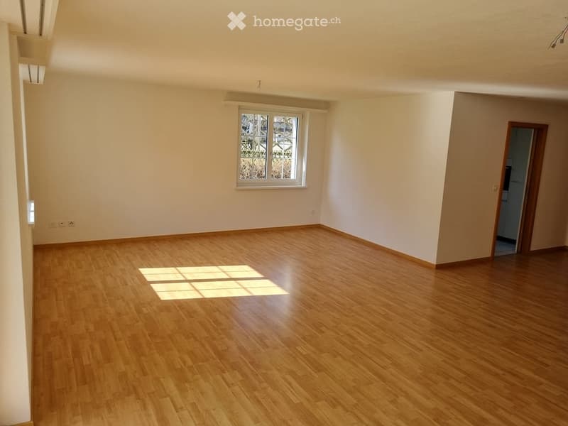 Completely empty room with wooden flooring and white walls. A single window with a grid pattern allows natural light to enter, and there is a doorway leading to another room.