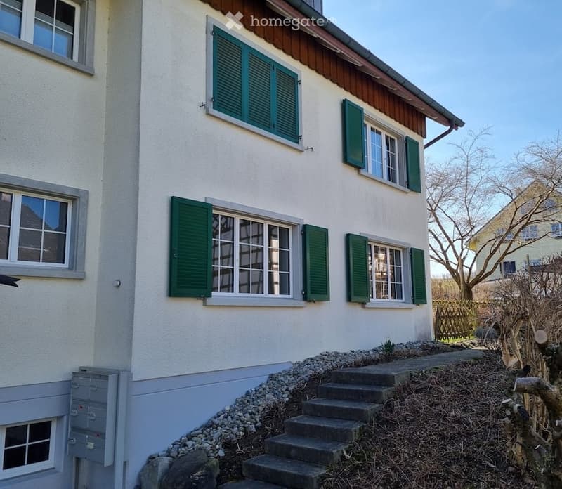 Two-story house with white siding, green shutters, and multiple windows. Stairs lead up to the entrance.