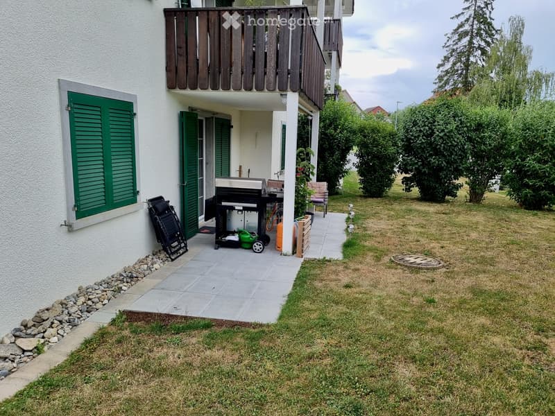 Garden area with a barbecue, a chair, and a garden cart, surrounded by green bushes and a paved pathway leading to the house.