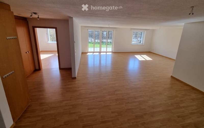 Empty room with wooden flooring, white walls, large windows, and a door leading to another room.