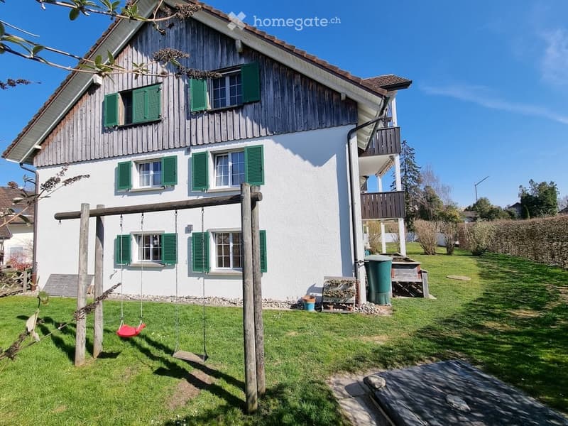 Two-story house with white walls, green shutters, and a wooden balcony. The front yard features a swing set and a small garden area.