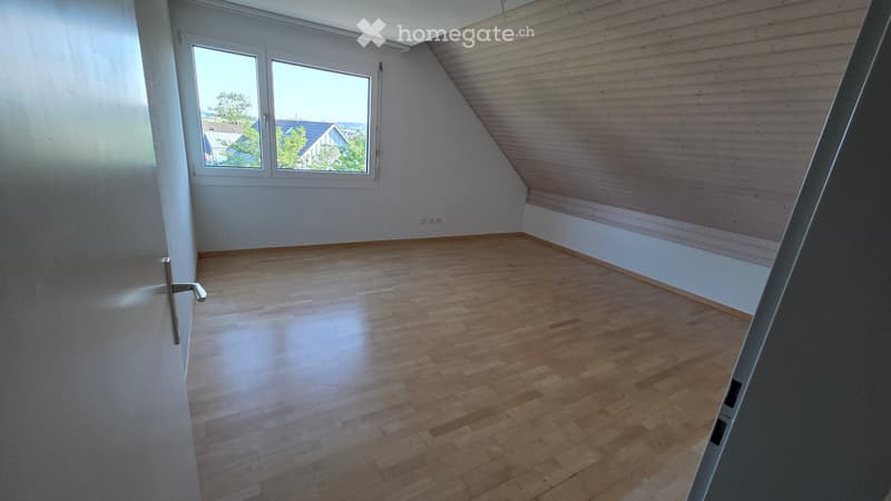 Empty attic room with wooden flooring, white walls, and large windows offering a view of the outside. The room features a slanted wooden ceiling and a door on the left side.