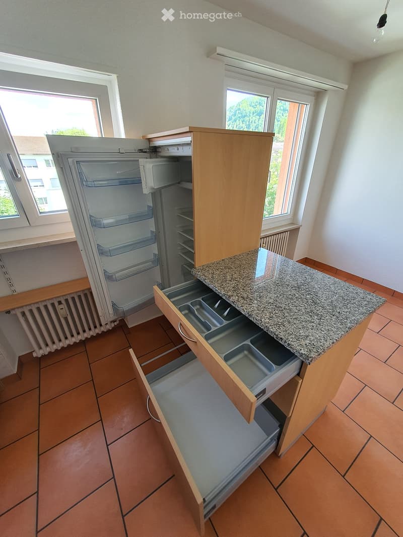 Modern kitchen with a granite countertop, wooden cabinets, and a refrigerator. Drawers and cabinets are visible, with a window providing natural light.