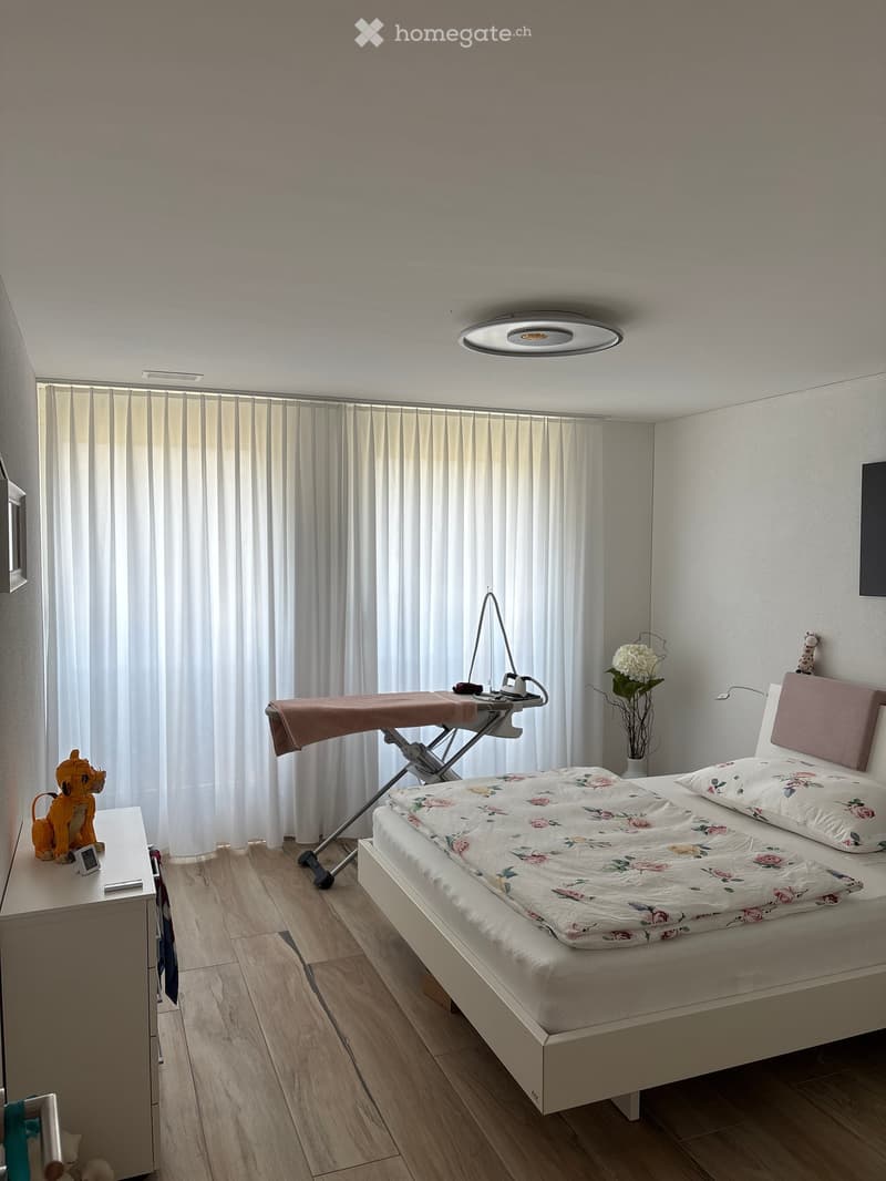 A well-lit bedroom with a white bed, floral quilt, nightstand, lamp, ironing board, vase with flowers, and decorative toys.