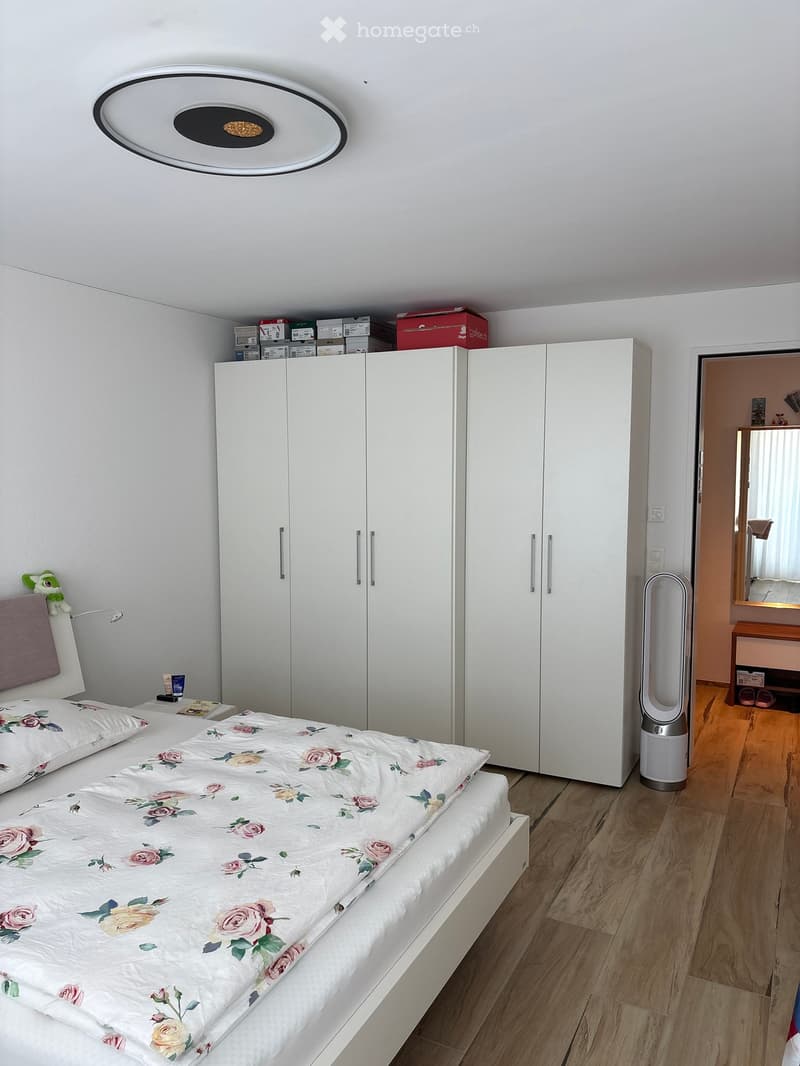 Modern bedroom with a white bed featuring a floral duvet, a large white wardrobe, a wall-mounted lamp, and a sleek fan near the doorway.