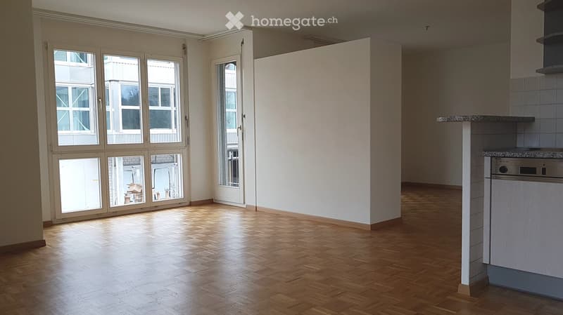 Empty room with wooden flooring, large windows, and a partially visible kitchen area with a refrigerator and countertop.