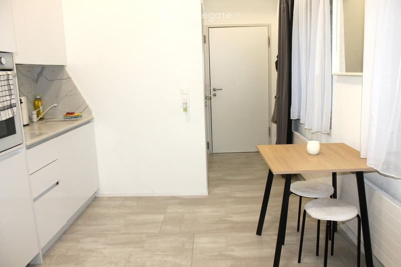 A modern, compact kitchen area with white cabinets, a small wooden table with two stools, and a sink area with a faucet and a bottle of dish soap. The space is well-lit with natural light coming from a window with white curtains.