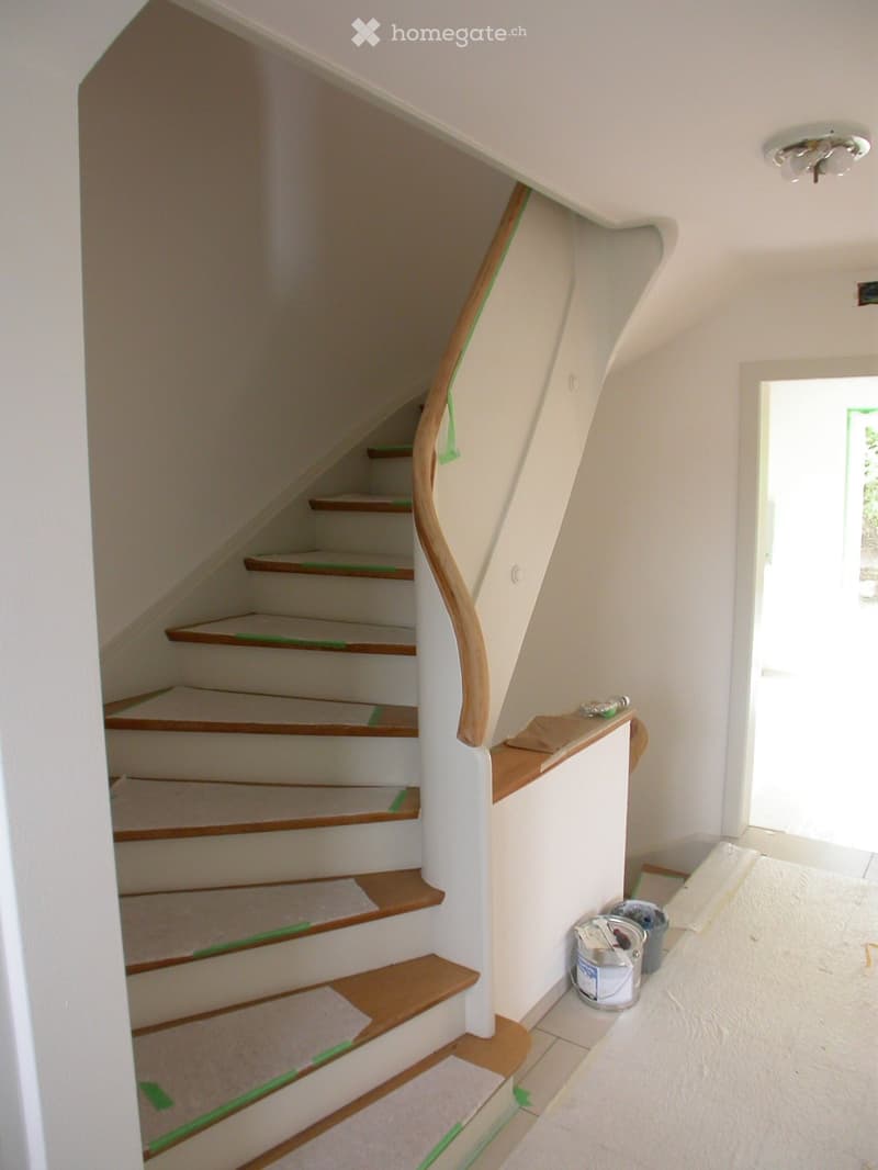 Staircase with wooden steps and a curved wooden handrail, white walls, and a small landing area with a window and construction materials.