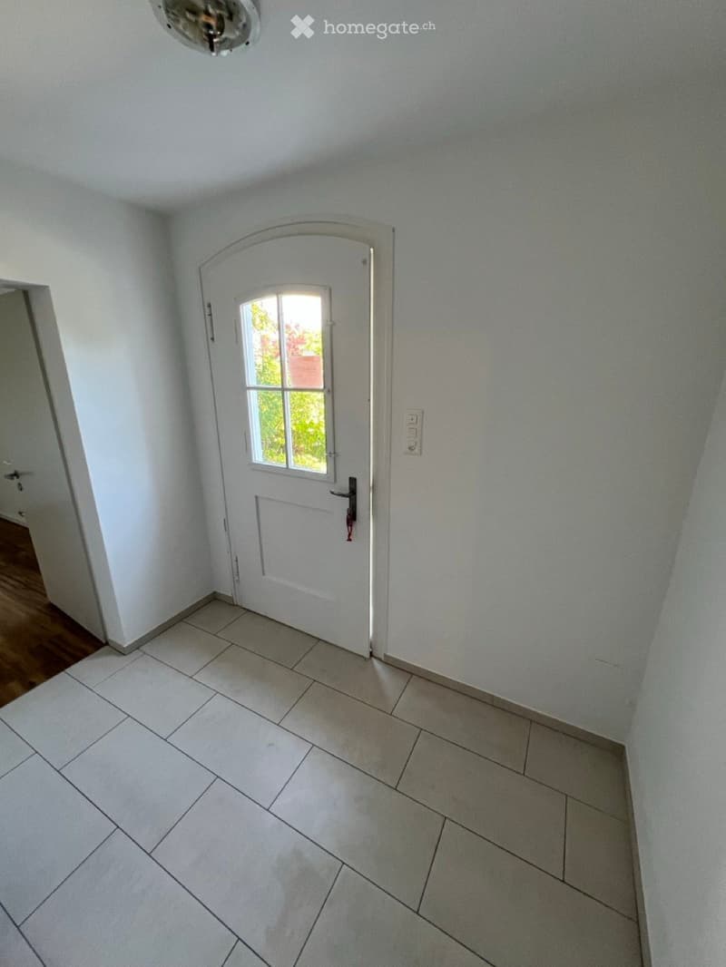 Empty hallway with white walls, tiled floor, and a white door with a window leading to another room.
