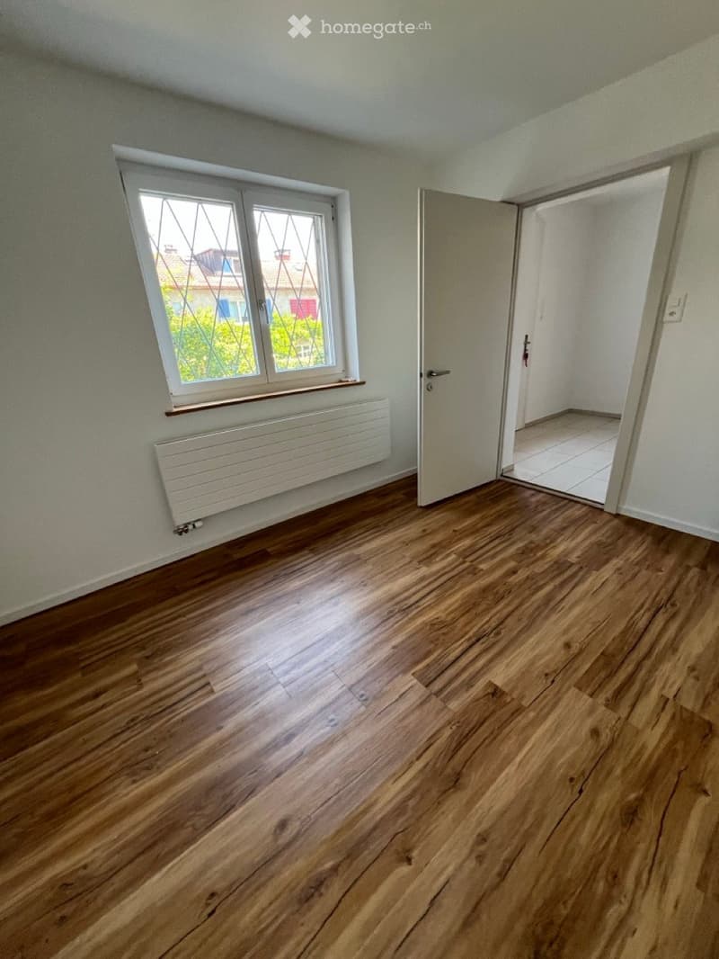 Empty room with wooden flooring, white walls, a window with frosted glass, a radiator, and an open door leading to another tiled room.